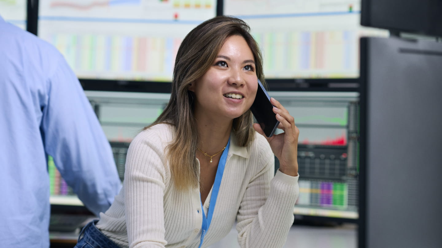 Person speaking on a phone in an office with multiple monitors displaying charts and data in the background
