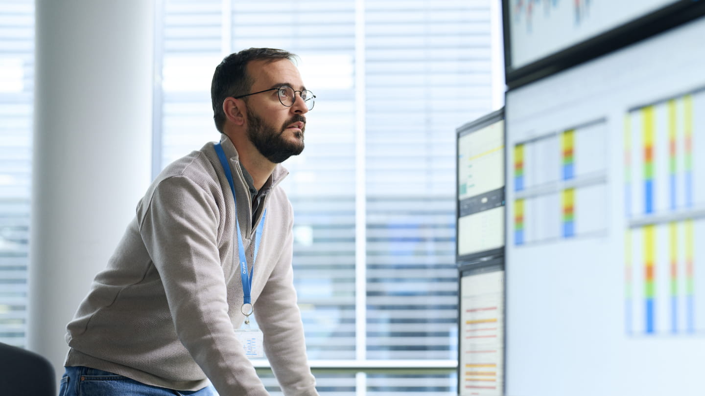 Person wearing an ID badge reviews data visualisations on multiple monitors in a control-room style office