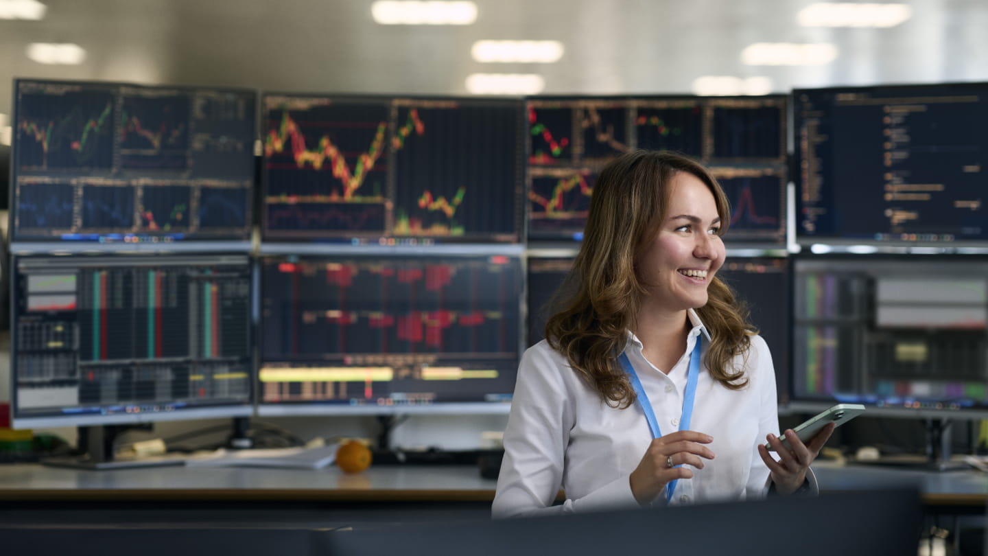 Person holding a smartphone in front of multiple computer monitors displaying market charts and trading data