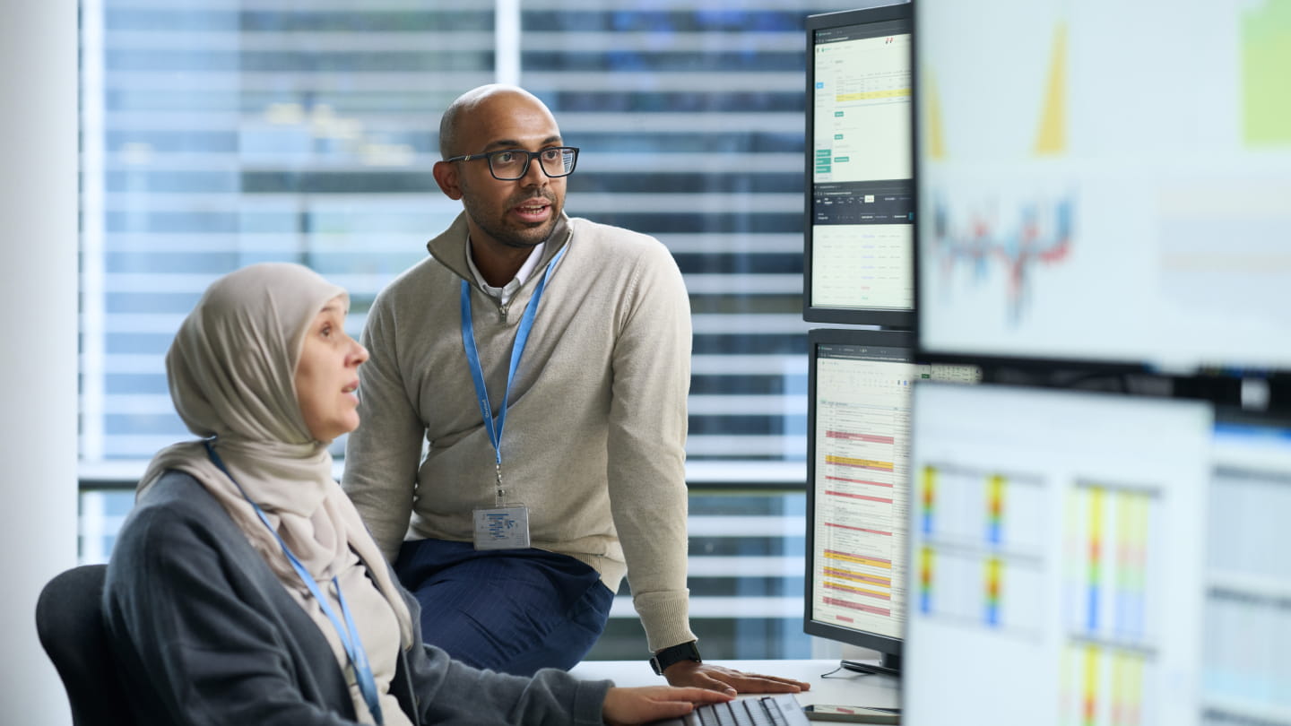 Two colleagues in an office reviewing data on multiple computer monitors, with charts and spreadsheets visible