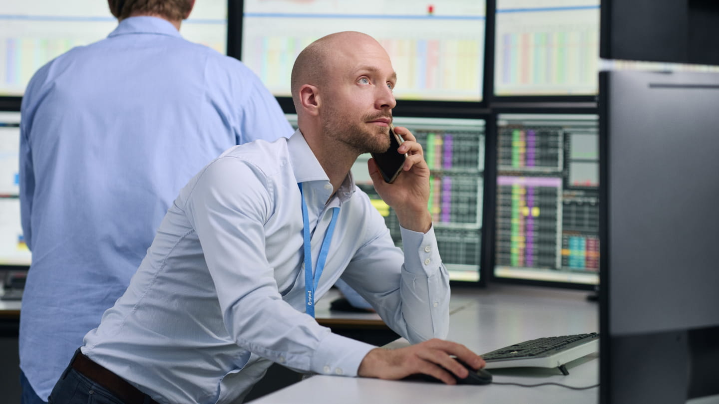 Person on a phone call at a desk, using a computer while multiple monitors behind display charts and data
