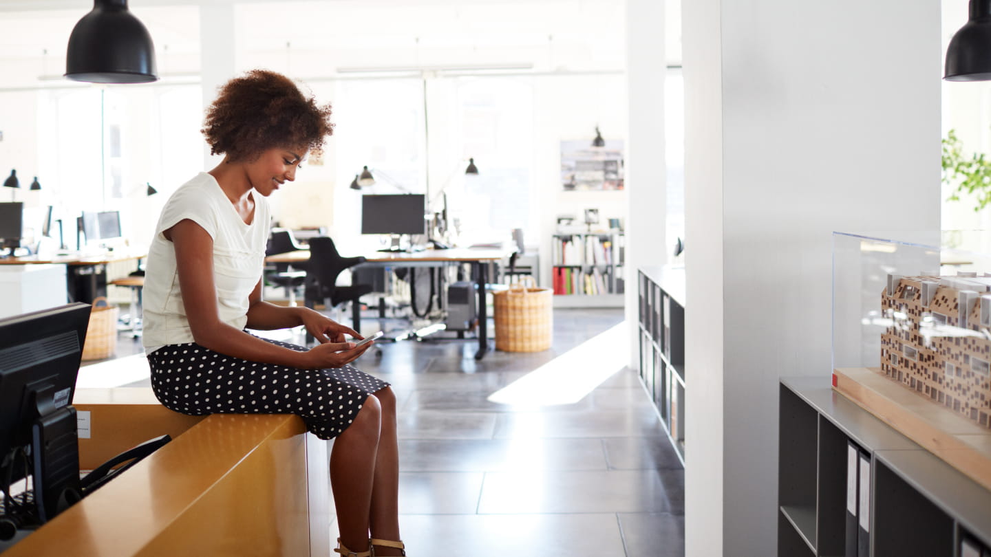 Person sitting on a counter in a bright open-plan office, looking at a smartphone