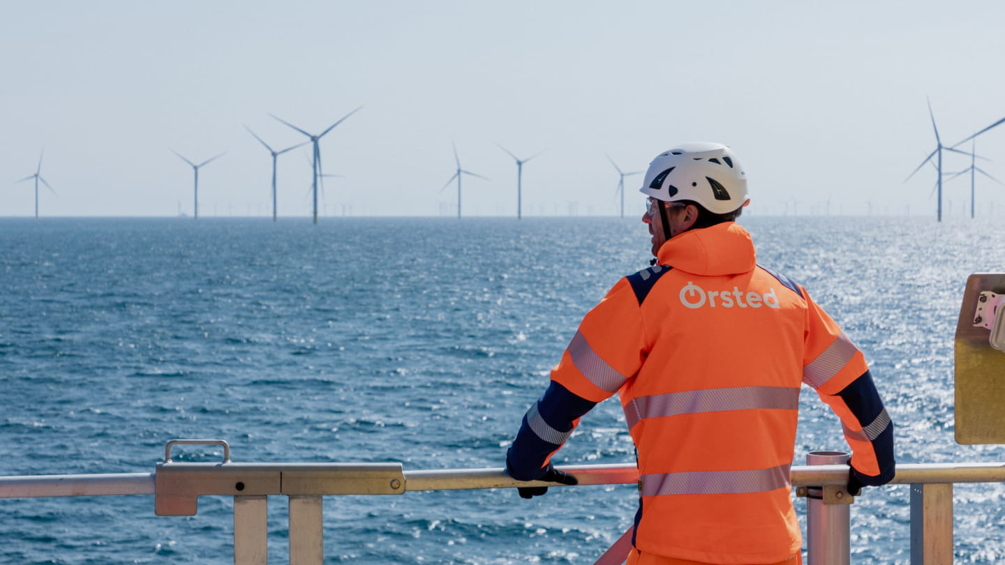 Ørsted technician in high-visibility jacket and helmet looking out over the sea toward an offshore wind farm