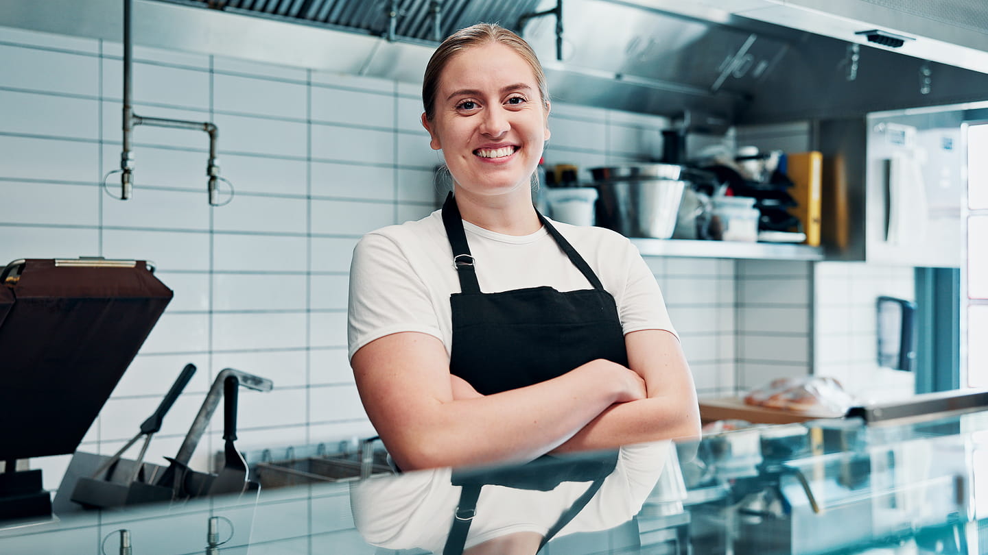 Worker in an apron standing with arms crossed behind a counter in a commercial kitchen