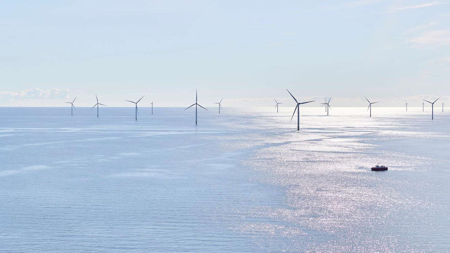 Anholt Wind Farm (ANH) consists of 111 wind turbines on a calm, sunlit sea beneath a pale blue sky; small service vessel at right.
