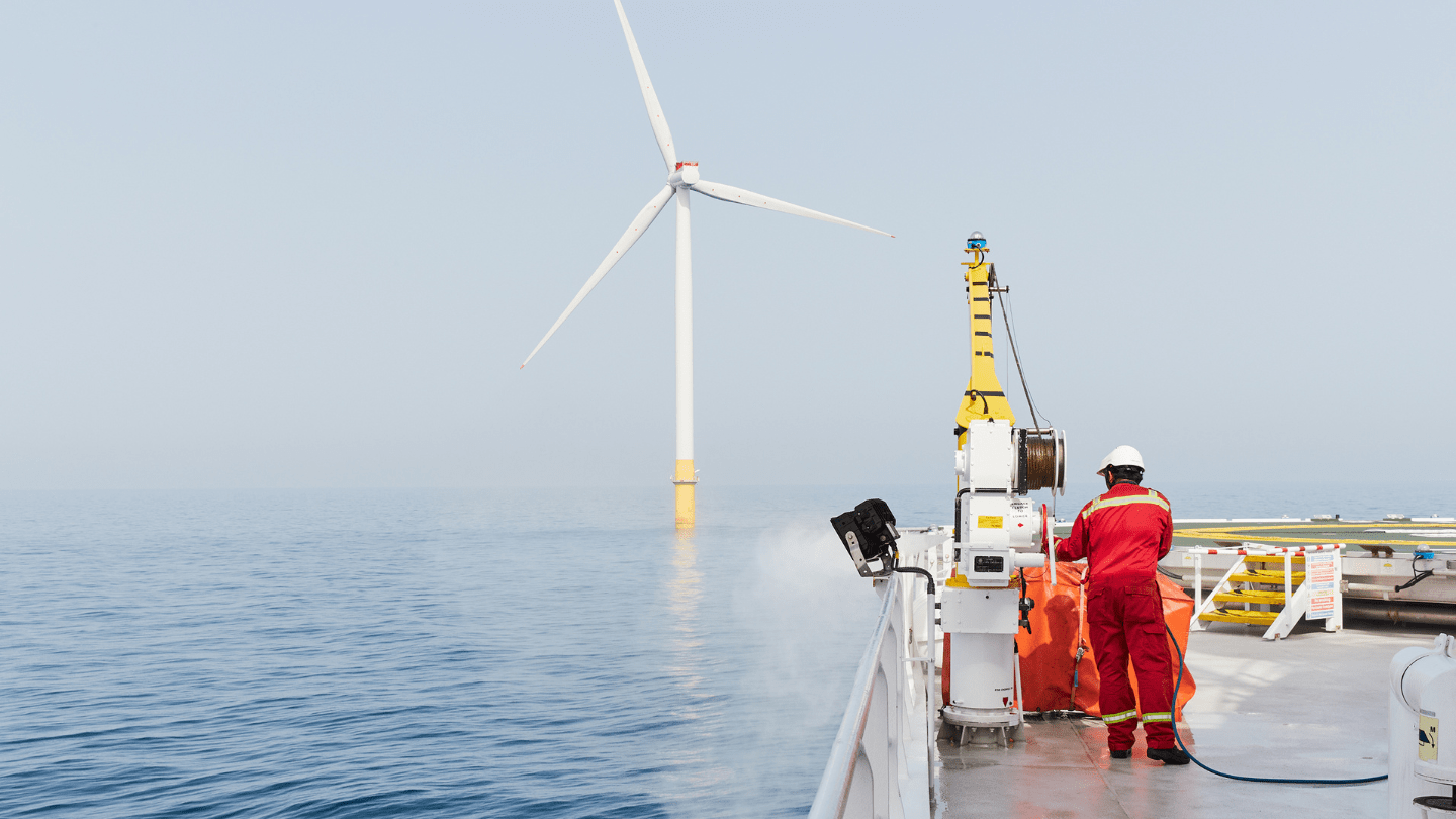 Offshore wind turbine seen from a service vessel; technician in red PPE on deck.