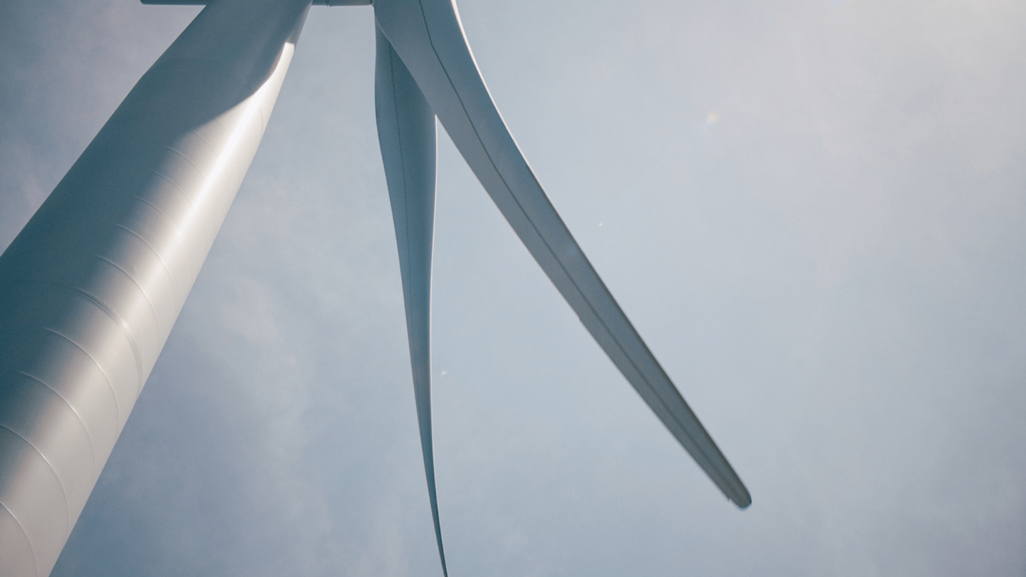Close-up, upward view of a wind turbine tower with two blades against a pale blue sky.