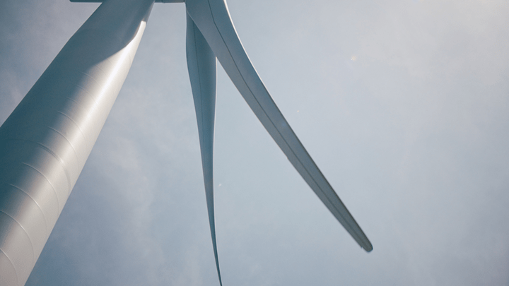 Close-up, upward view of a wind turbine tower with two blades against a pale blue sky.