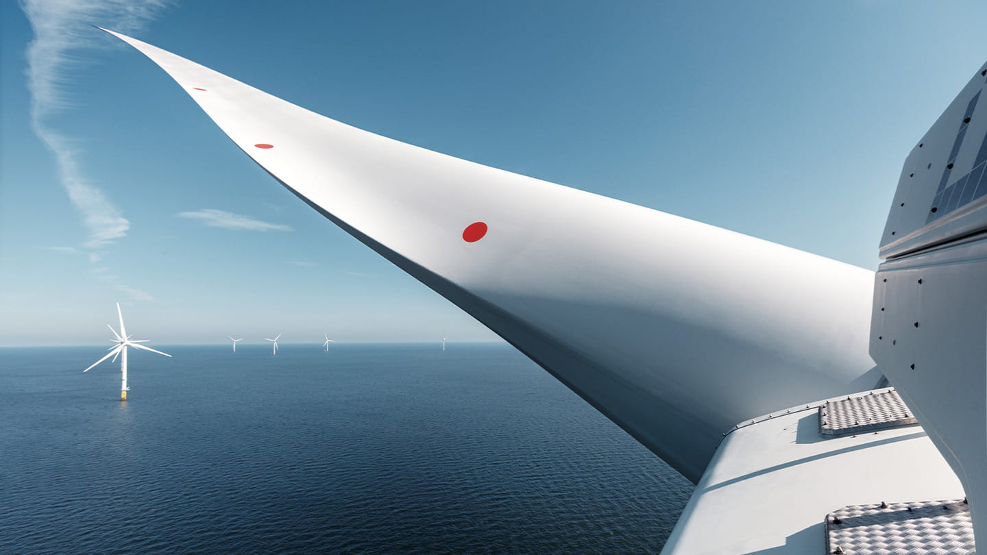 Close-up view from a wind turbine showing a white blade over the sea, with several offshore wind turbines in the distance under a clear blue sky.