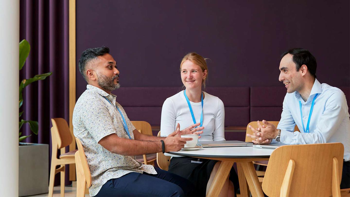 Three colleagues with lanyards chat at a round cafe table with notebooks and coffee.
