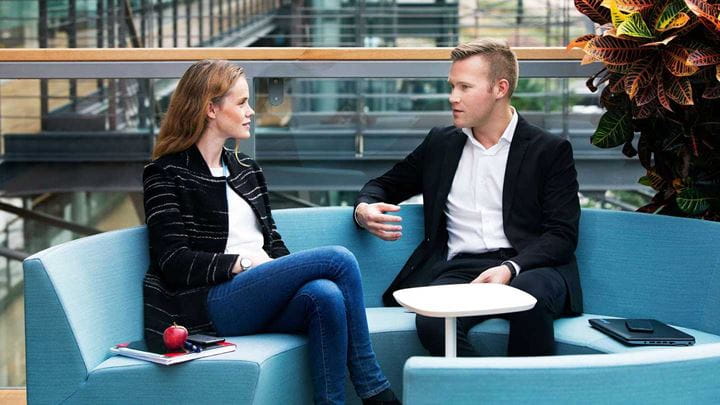 A man and a woman sitting on a blue circular sofa while engaging in conversation.