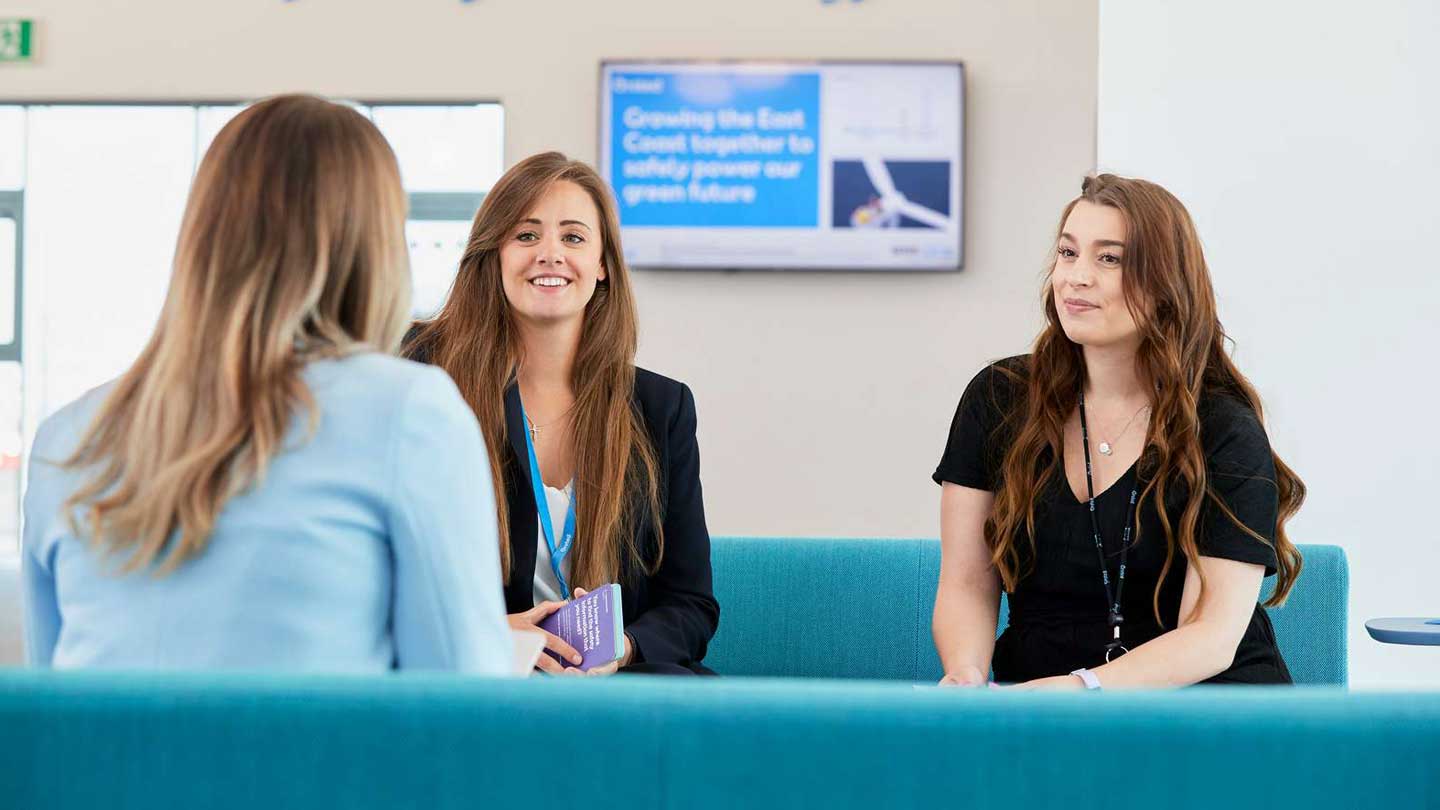 Three female Ørsted employees in office attire sitting on a blue sofa while talking.