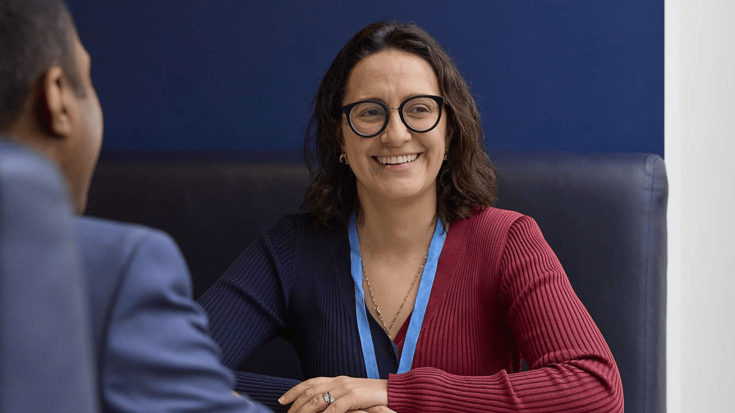 Portrait of employee in London Office. Sitting at a table, smiling, talking, dark blue wall.