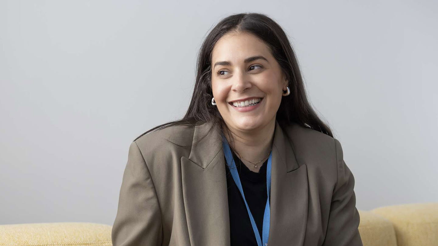 Person with long dark hair sitting on a yellow sofa, wearing a taupe blazer over a black top and blue lanyard in a plain office setting.