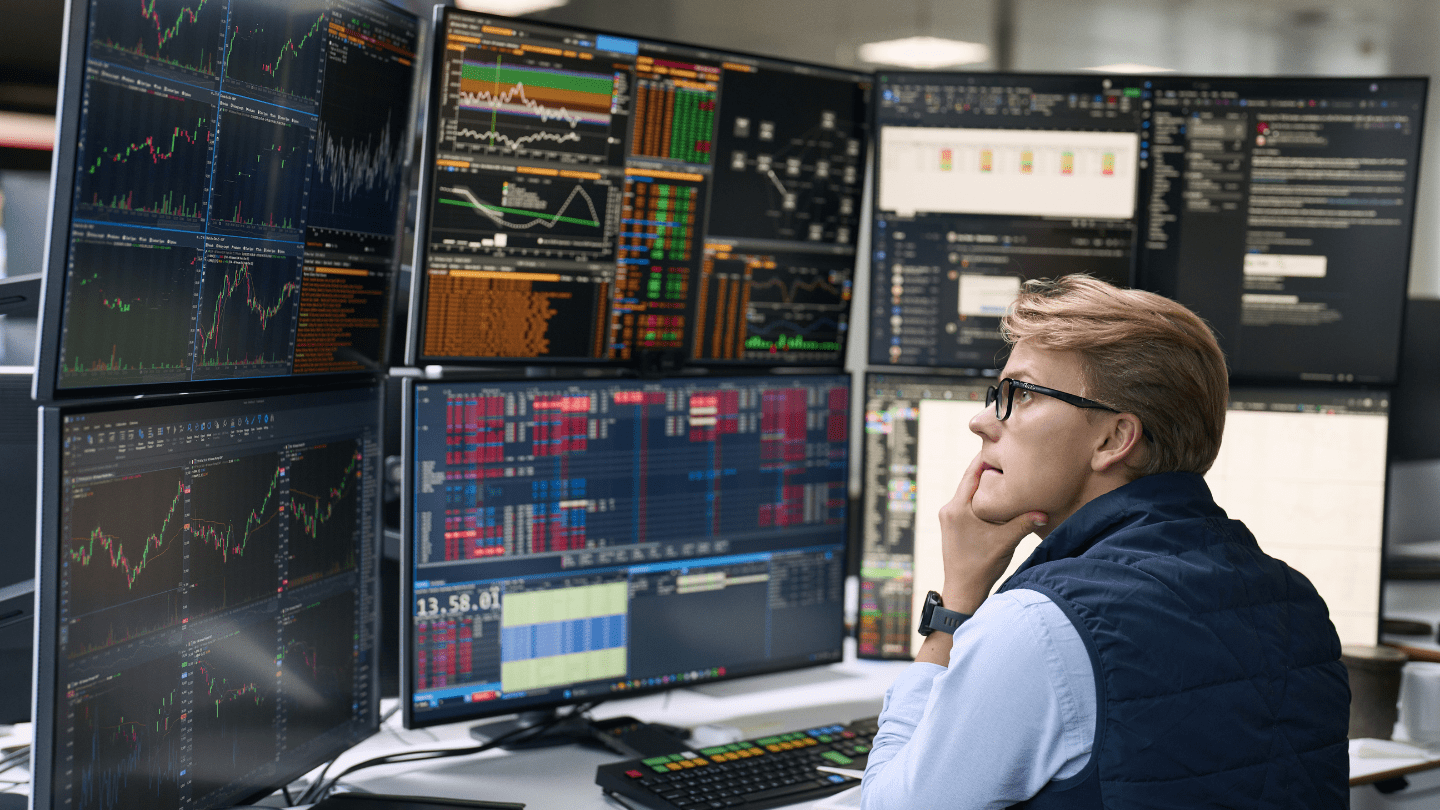 Employee seated at a trading desk, analyzing a wall of large monitors filled with colorful financial charts, order books, dashboards, and chat windows in a modern office setting.