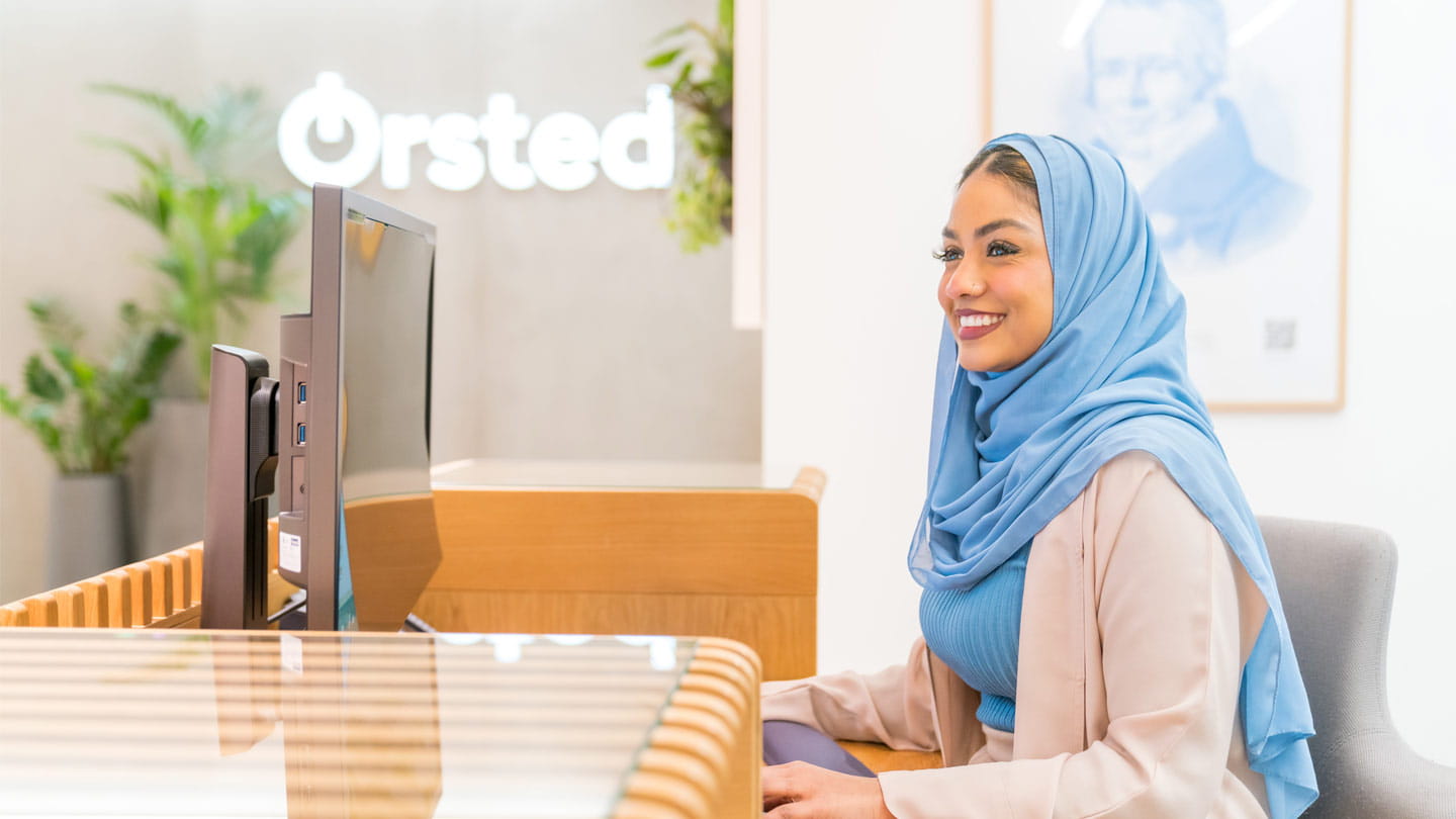 Receptionist wearing a blue hijab works at a computer behind a wooden desk in a bright office, with the Ørsted logo and indoor plants in the background.