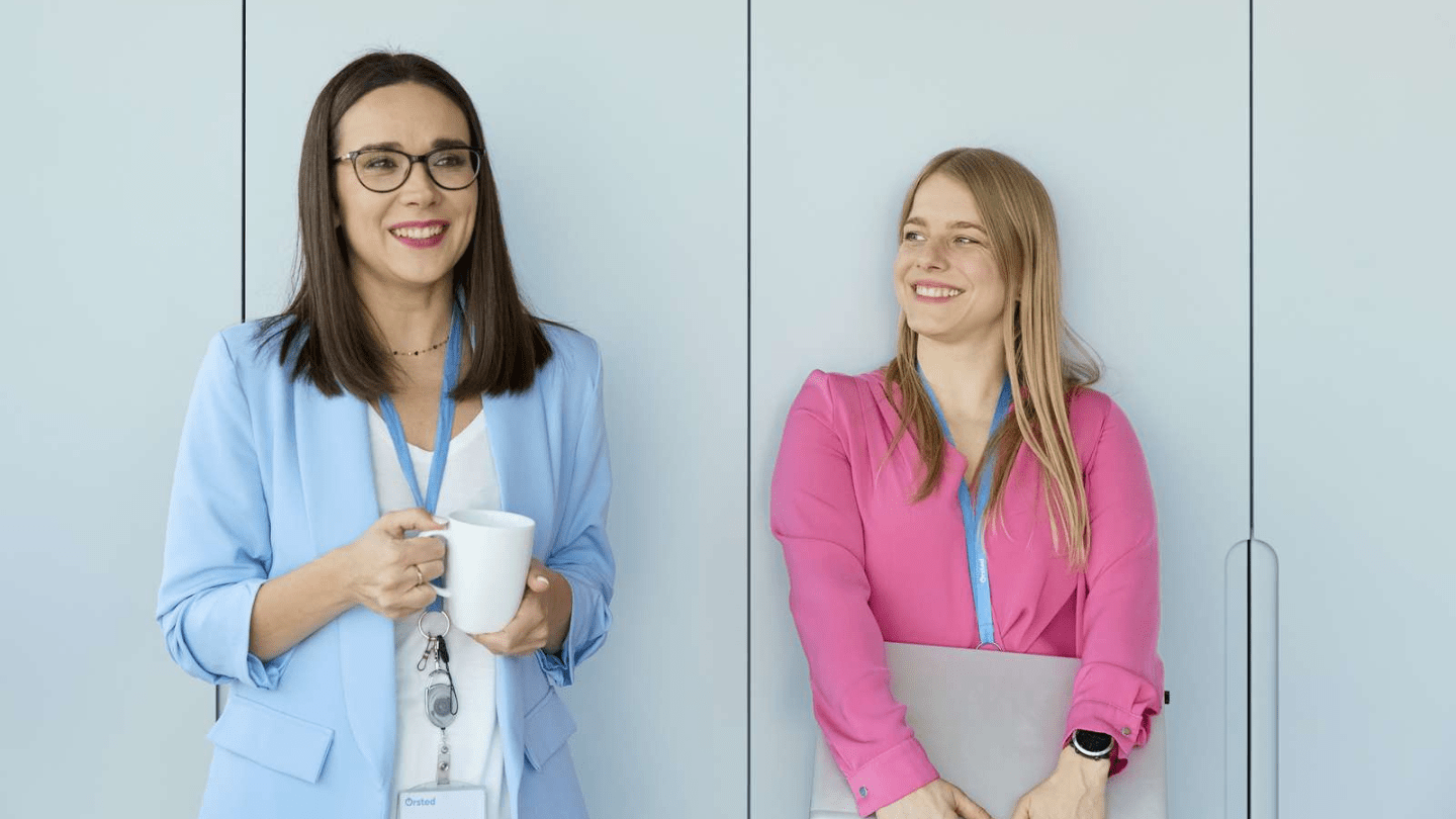 Warsaw office. Two office colleagues stand against a light gray wall; one in a light blue blazer holds a white mug and wears a lanyard with an ID badge, while the other in a pink blouse wears a smartwatch and lanyard and holds a closed laptop