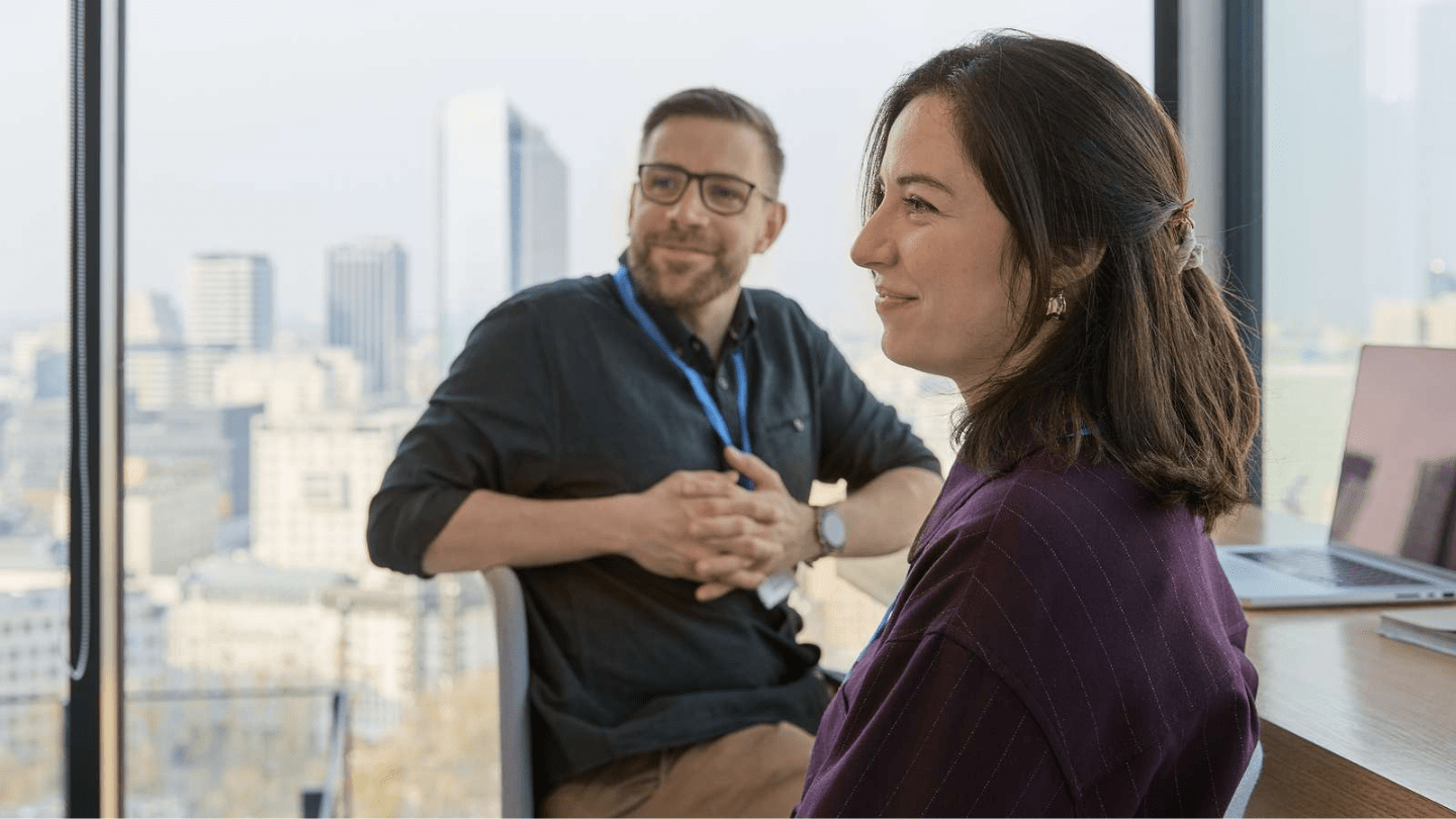 Poland office. Two colleagues in an office lounge by floor-to-ceiling windows talk, both wearing blue lanyards, with a city skyline in the background and an open laptop and notebook on the table.