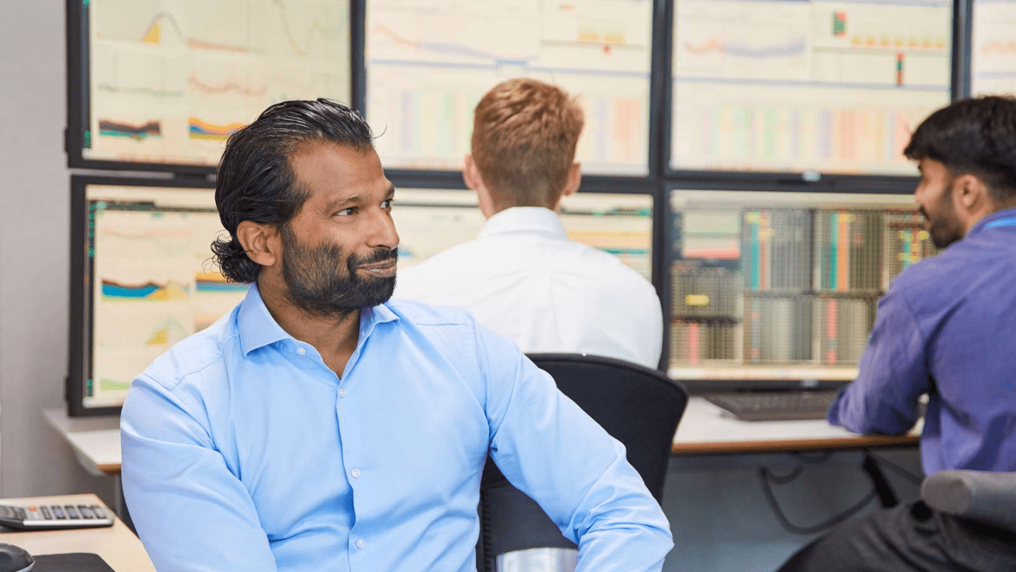 Colleagues in a trading or operations control room, with multi-screen dashboards displaying charts and data; a person in a blue shirt sits in the foreground while others work at their stations.