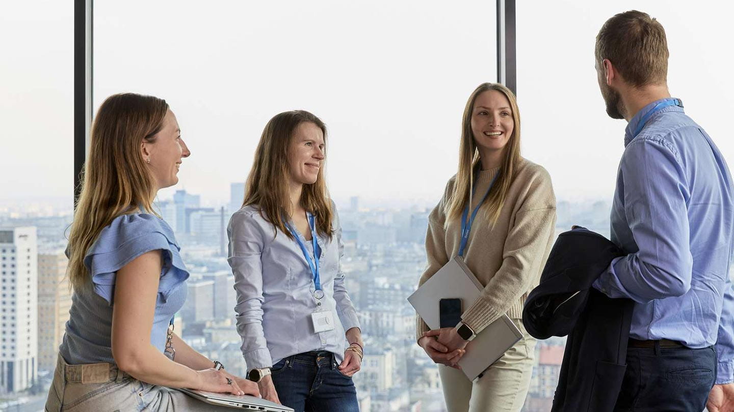 Four colleagues discuss by a tall window with city skyline, holding notebooks and tablets.