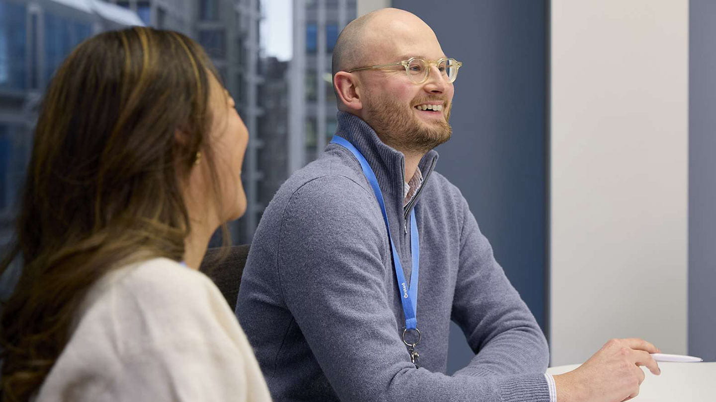 Two colleagues discuss at a conference table; one holds a pen, lanyards visible, city buildings outside.
