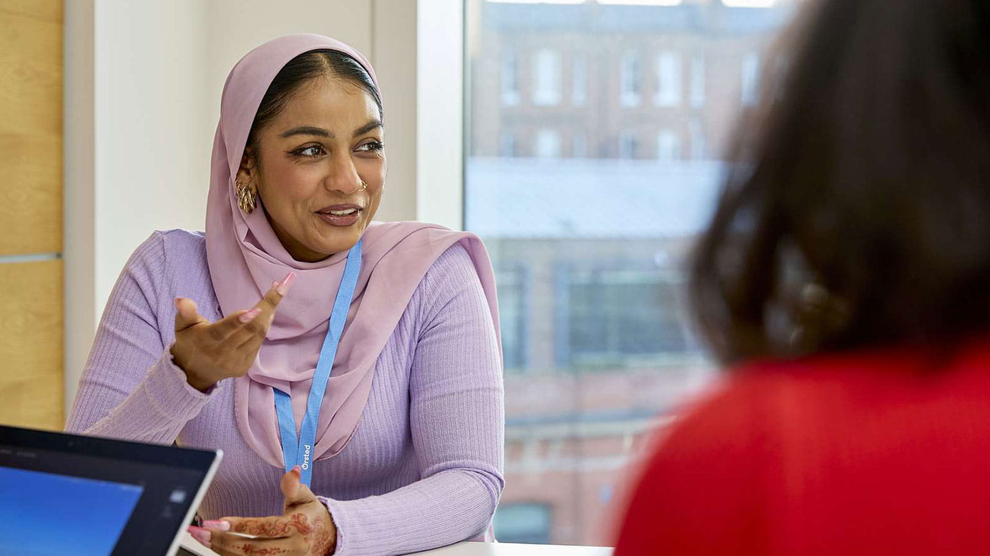 Person in a pink headscarf gestures in an office meeting; laptop and city skyline by the window.