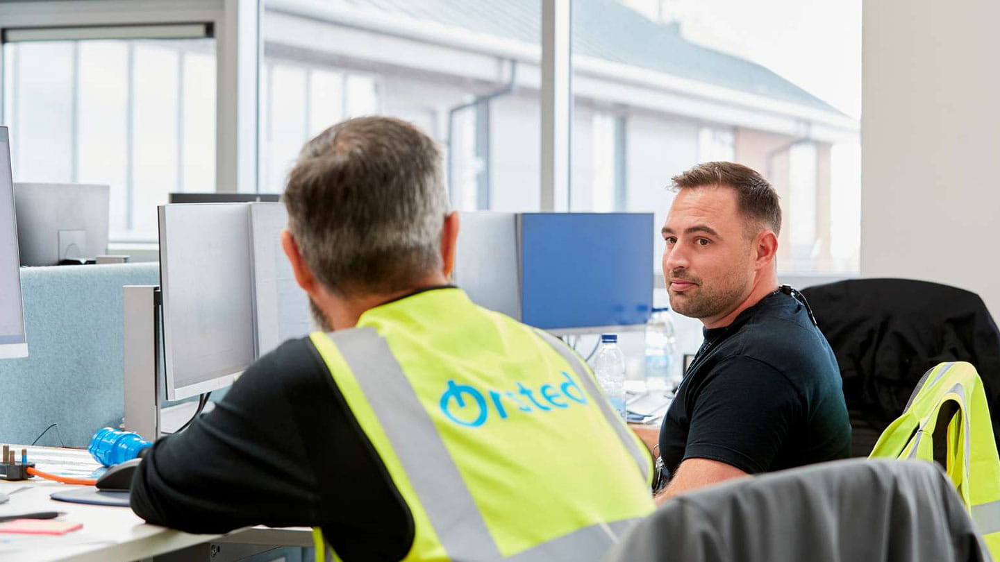 Orsted colleagues, one in branded high-vis vest, collaborate at multi-screen workstations. 