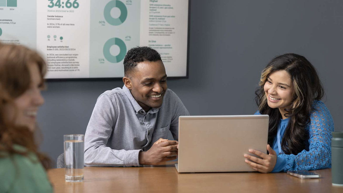 Coworkers collaborate at a laptop in a meeting room with data charts on the wall.