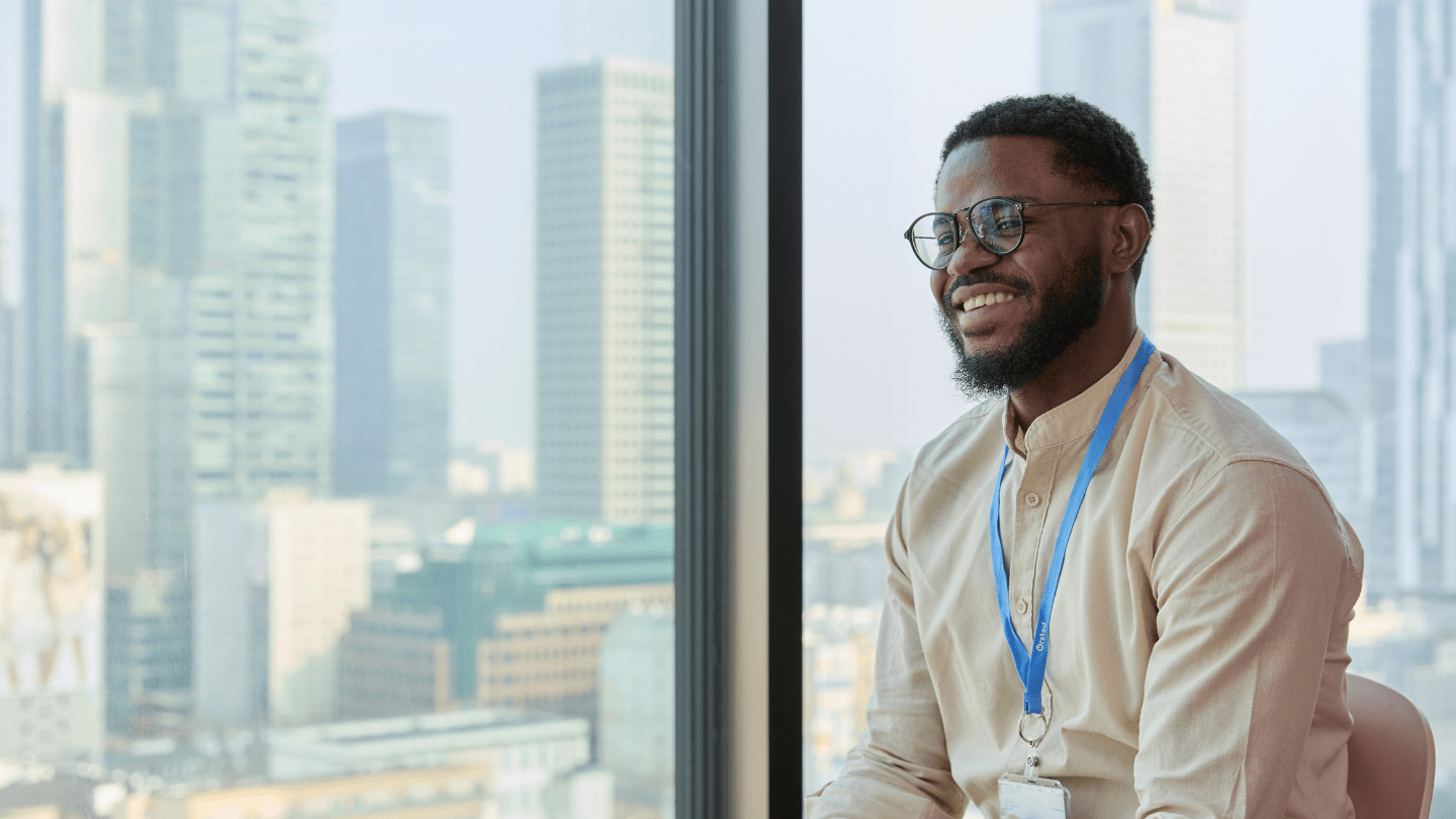Person with blue lanyard in beige shirt sits by a large window overlooking a modern city skyline.
