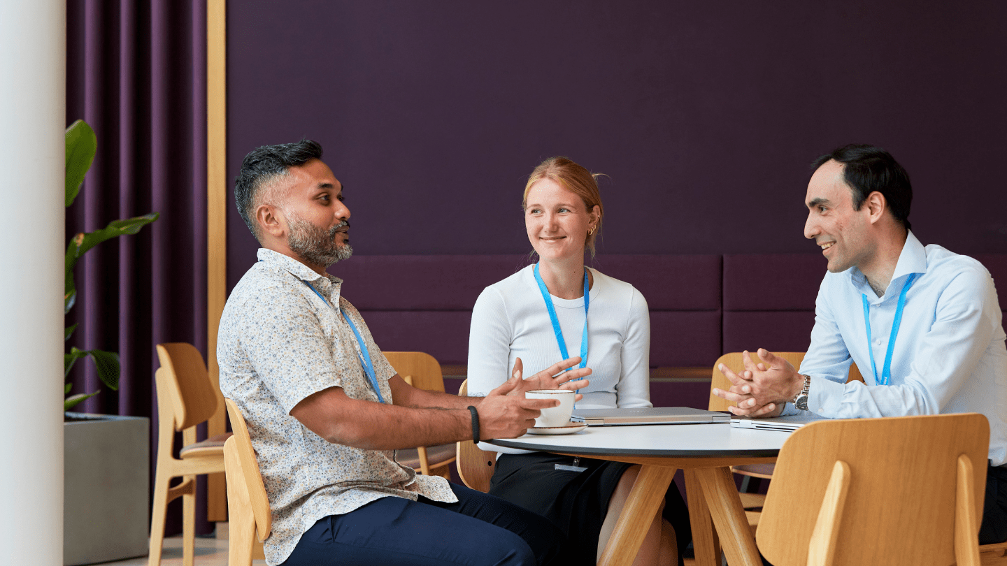 Gentofte office. Three colleagues wearing blue lanyards sit around a small round table, talking with a laptop and coffee cups on the table.