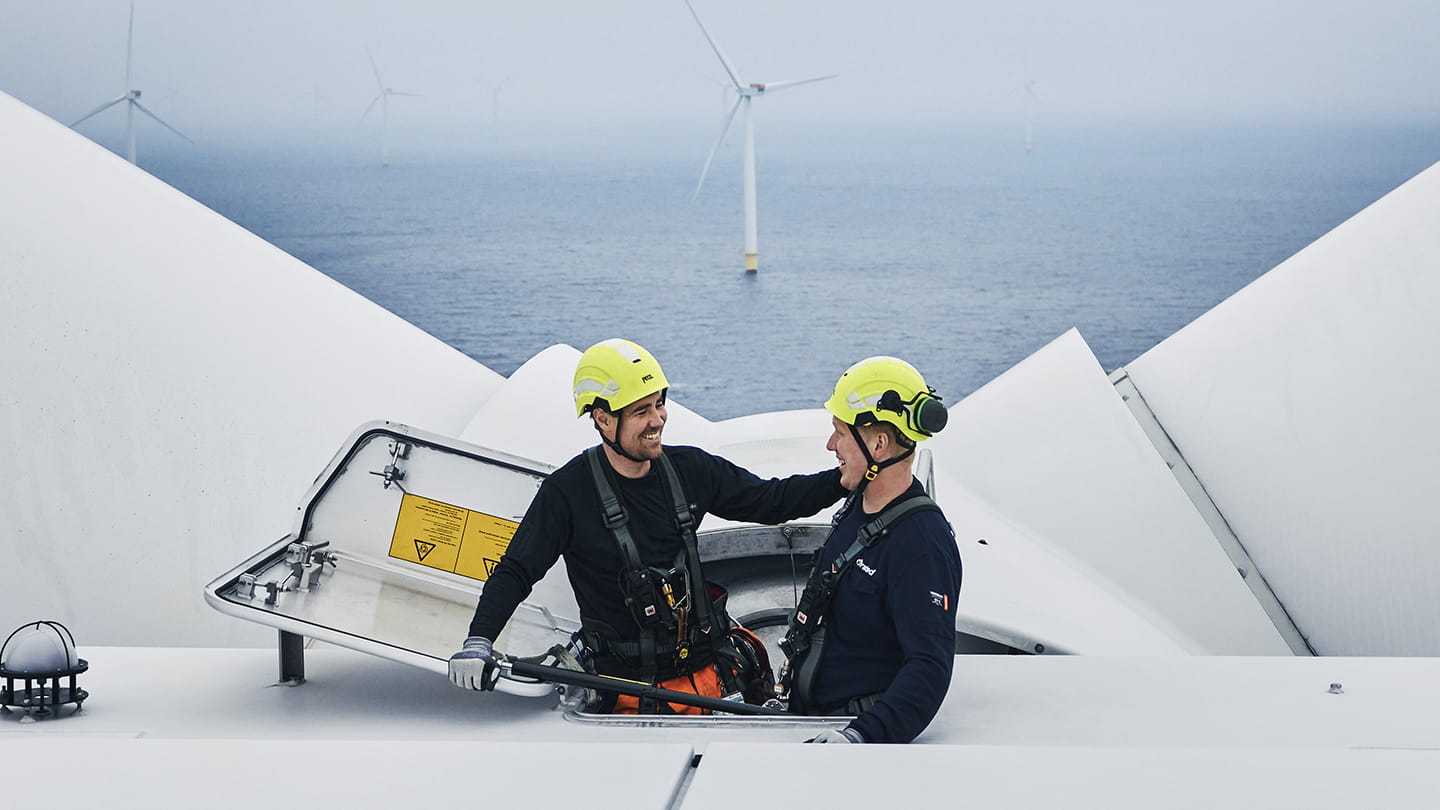Two wind turbine technicians in safety helmets and harnesses stand on top of a turbine nacelle with the sea and offshore wind turbines in the background.