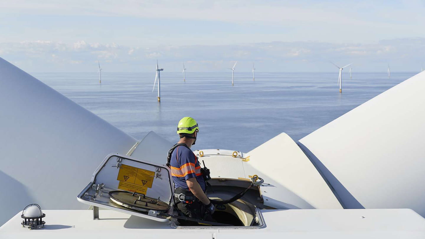 Technician in safety gear stands on the nacelle of an offshore wind turbine beside an open hatch, looking over a calm sea with distant turbines.