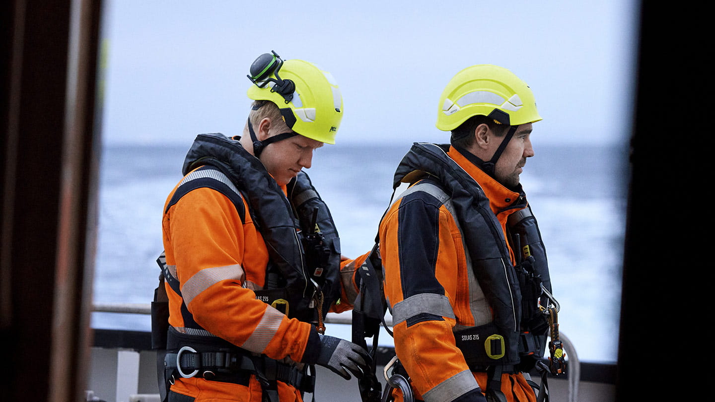  Two offshore wind technicians in orange and black survival suits and yellow helmets stand on a vessel, looking out over the sea under a grey sky.