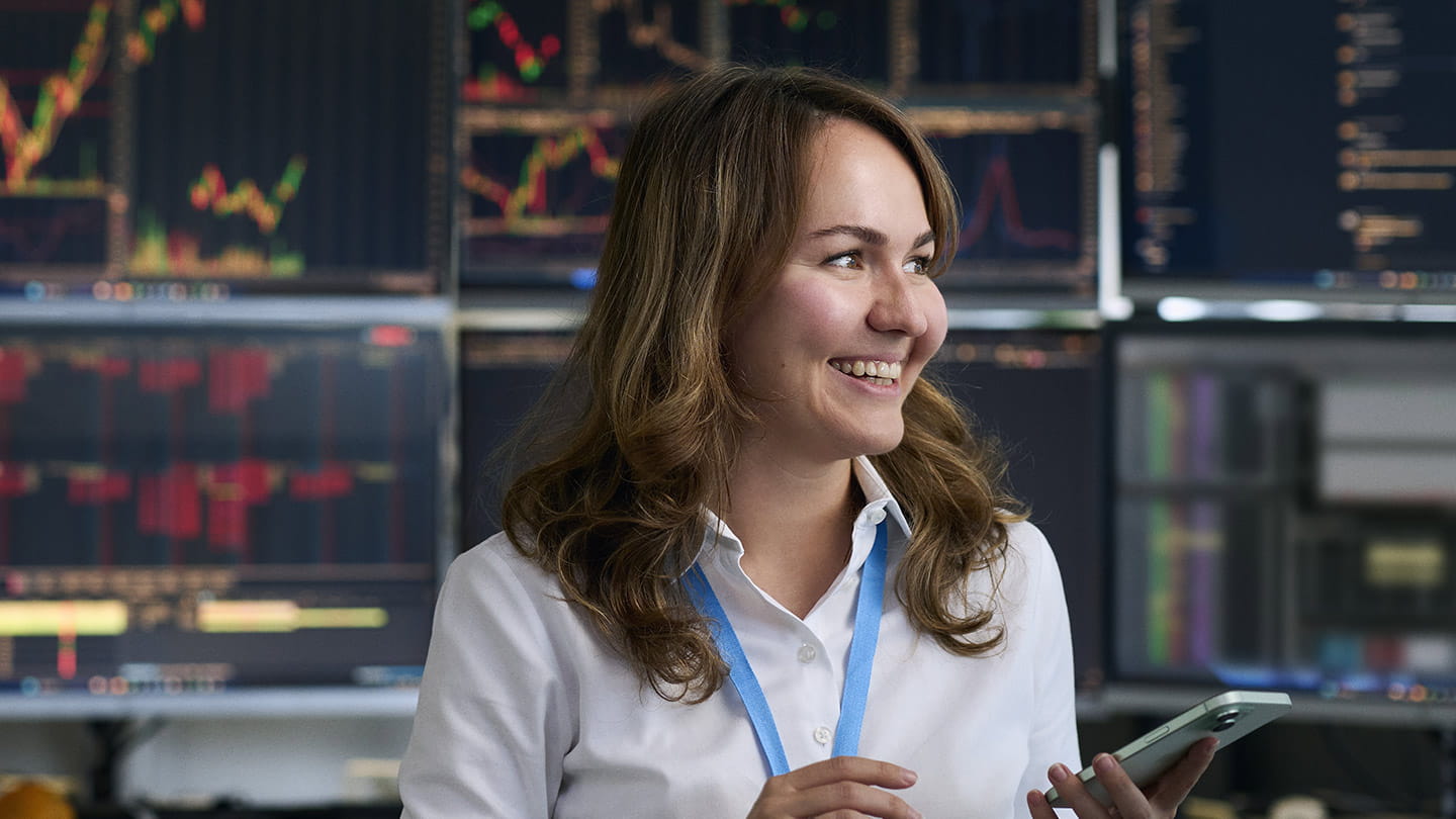 Person wearing a white blouse and blue lanyard, holding a tablet in a control-room setting with multiple data and chart screens in the background.