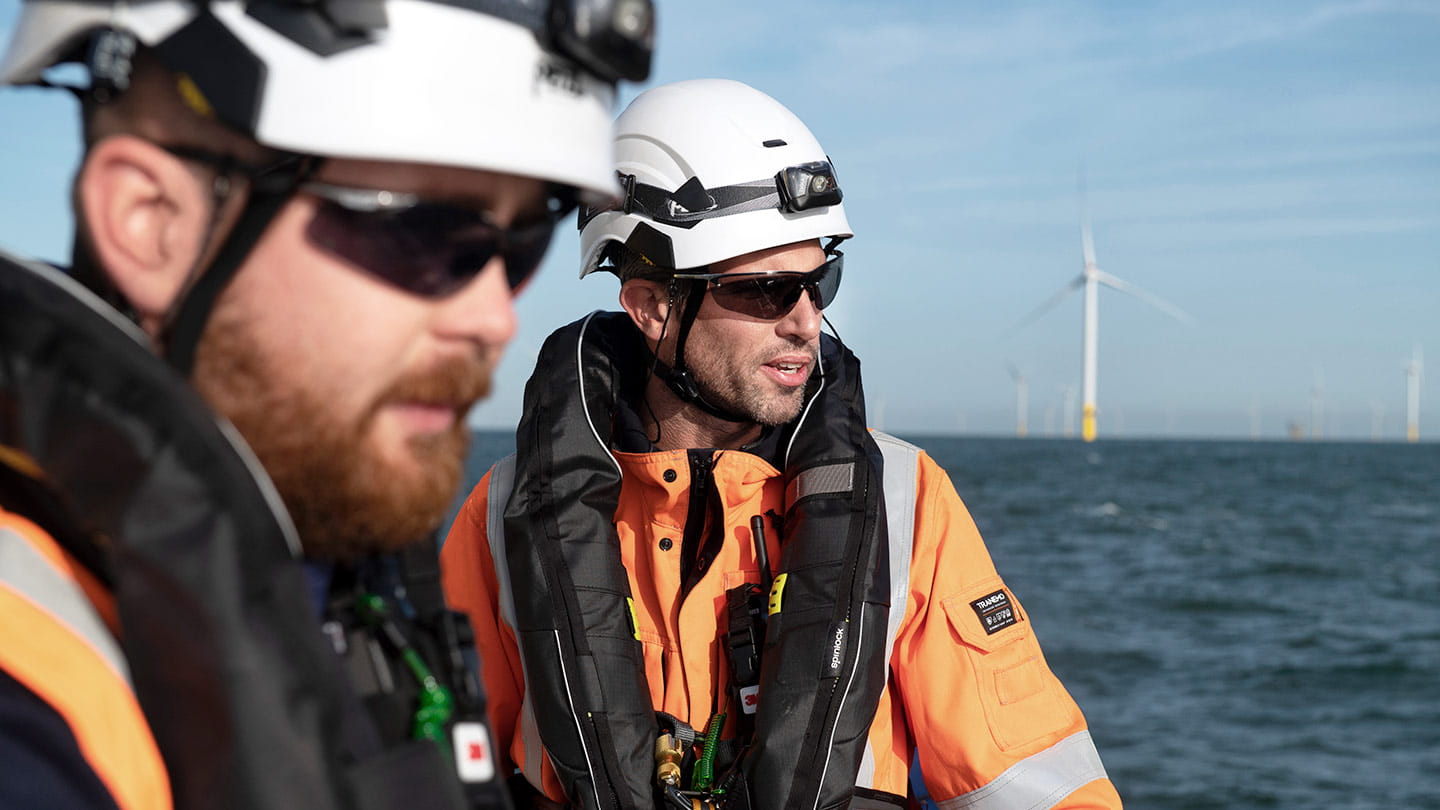  Two offshore workers in safety gear (helmets, life vests, and orange high-visibility jackets) on a vessel at sea, looking toward distant wind turbines.