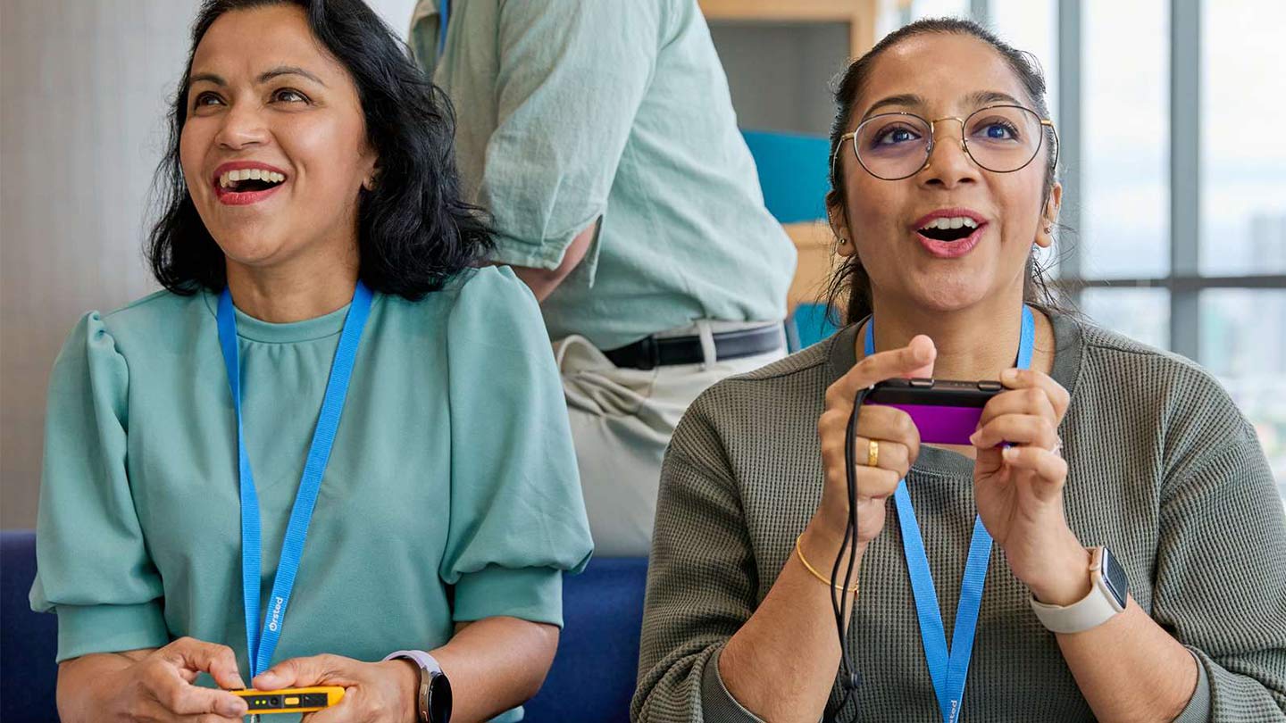 Two colleagues sit on a blue sofa in a modern office, each using small handheld gaming devices with wired controllers and wearing lanyards and smartwatches; another colleague stands behind near a table, with large windows and a city skyline in the background.