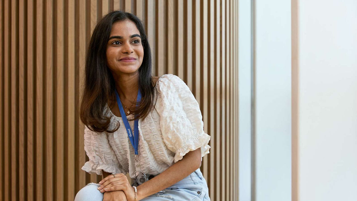 A smiling woman with shoulder-length dark hair sits on a yellow chair in a modern office with vertical wooden slat walls, wearing a white textured puff-sleeve blouse, light-wash jeans, and a blue Ørsted lanyard, her hands resting on her knee.