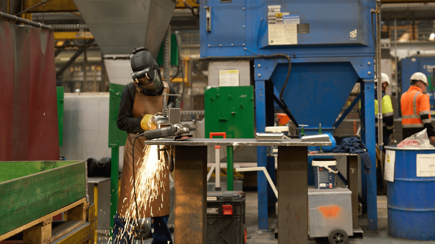 Industrial workshop scene: a worker in protective helmet, face shield, gloves, and leather apron uses an angle grinder on a metal piece, sending bright sparks, with large blue machinery, barrels, and other high-visibility–clad workers in the background.