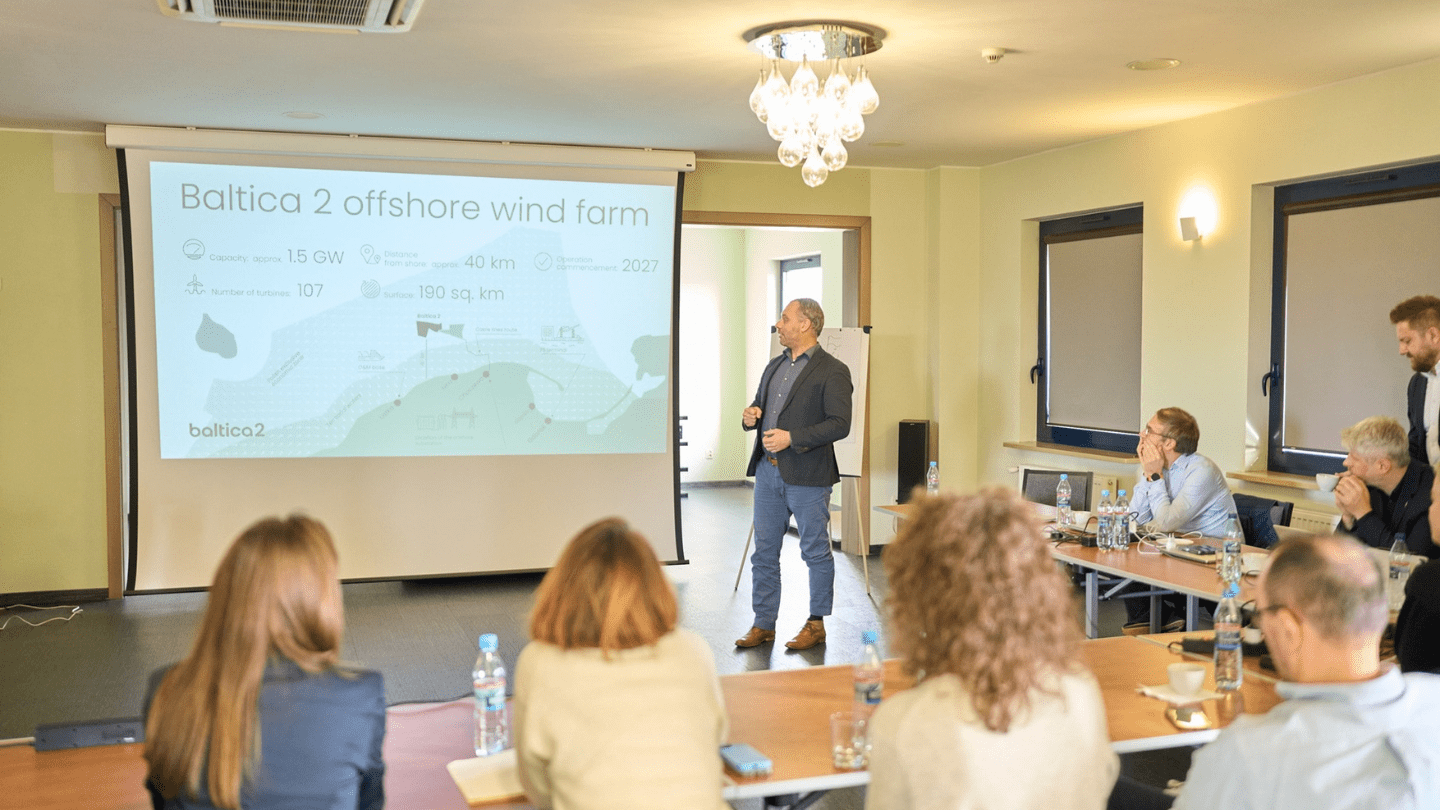 Meeting room presentation: a person stands beside a projector screen titled “Baltica 2 offshore wind farm,” addressing attendees seated at tables with notebooks, laptops, and water bottles. The slide shows a map and key figures—capacity approx. 1.5 GW, distance from shore approx. 40 km, operation commencement 2027, number of turbines 107, and surface 190 sq. km—under ceiling lights and near covered windows and a flipchart.