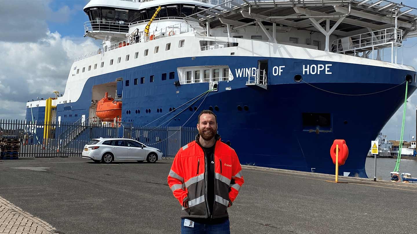 Person wearing a high-visibility jacket standing on a quay in front of the offshore support vessel “Wind of Hope” at a port.