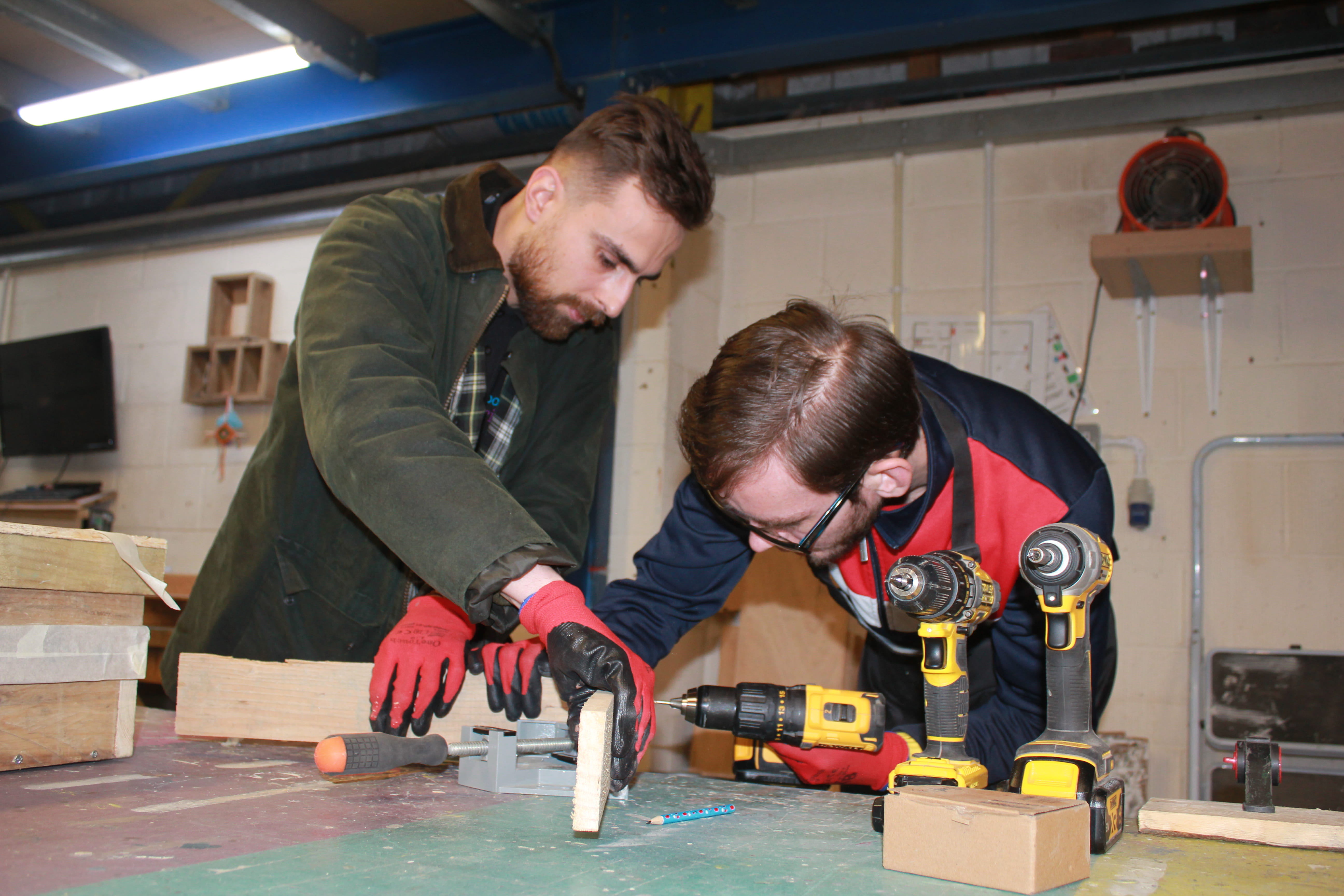 Two people in a workshop with one person holding a drill while the other holds the wood joint in place.