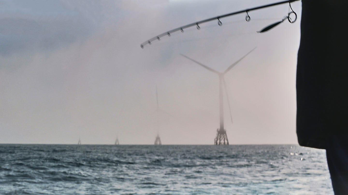 Close-up of a fishing rod held by Hank Hewitt, pointing toward the five turbines of Ørsted's Block Island Wind Farm.