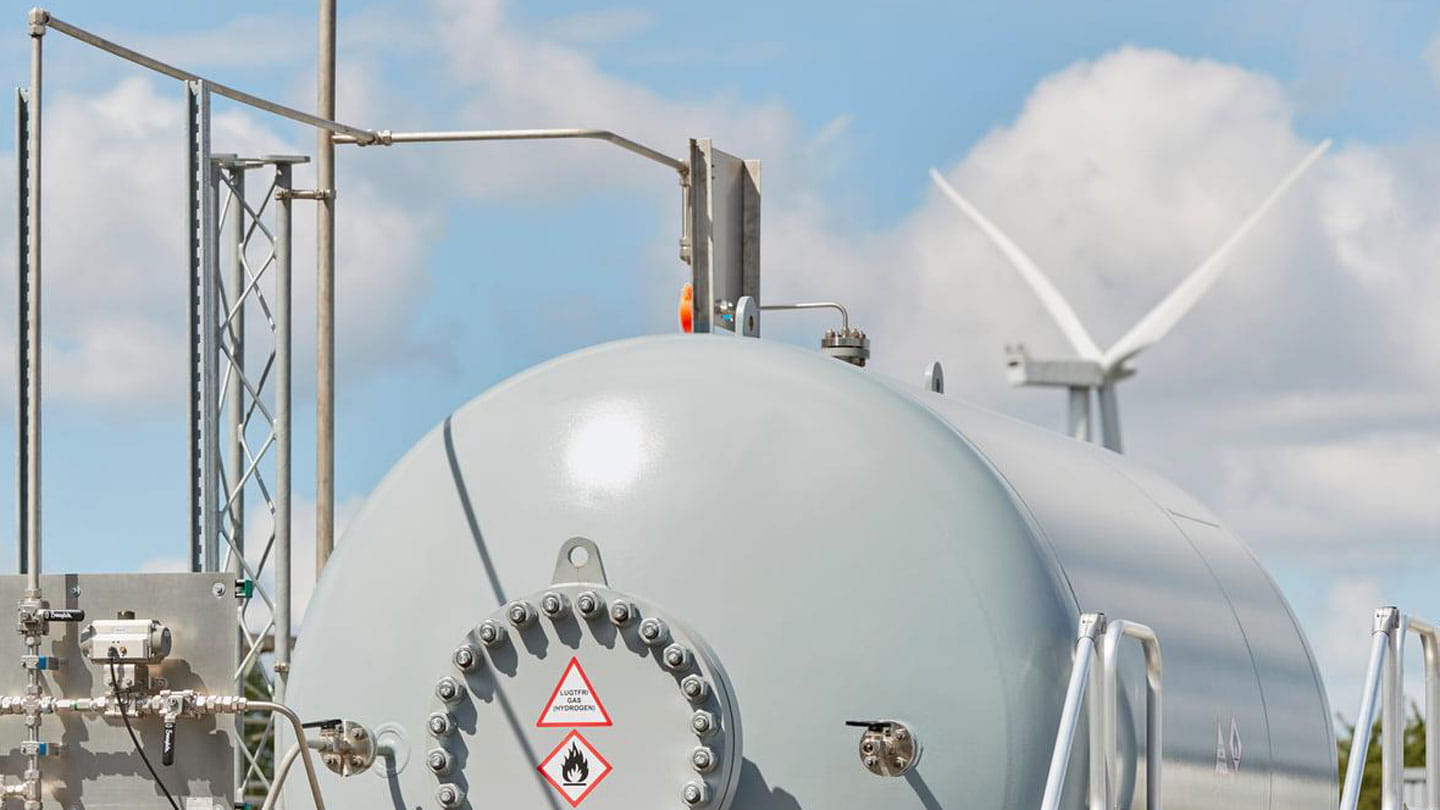 A large, gray hydrogen storage tank with a wind turbine in the background shows how Ørsted's Power-to-X facility work.